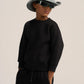 A child model wearing a black crewneck sweatshirt and black sweatpants, styled with a gray bucket hat and black sunglasses, against a plain light beige background.
