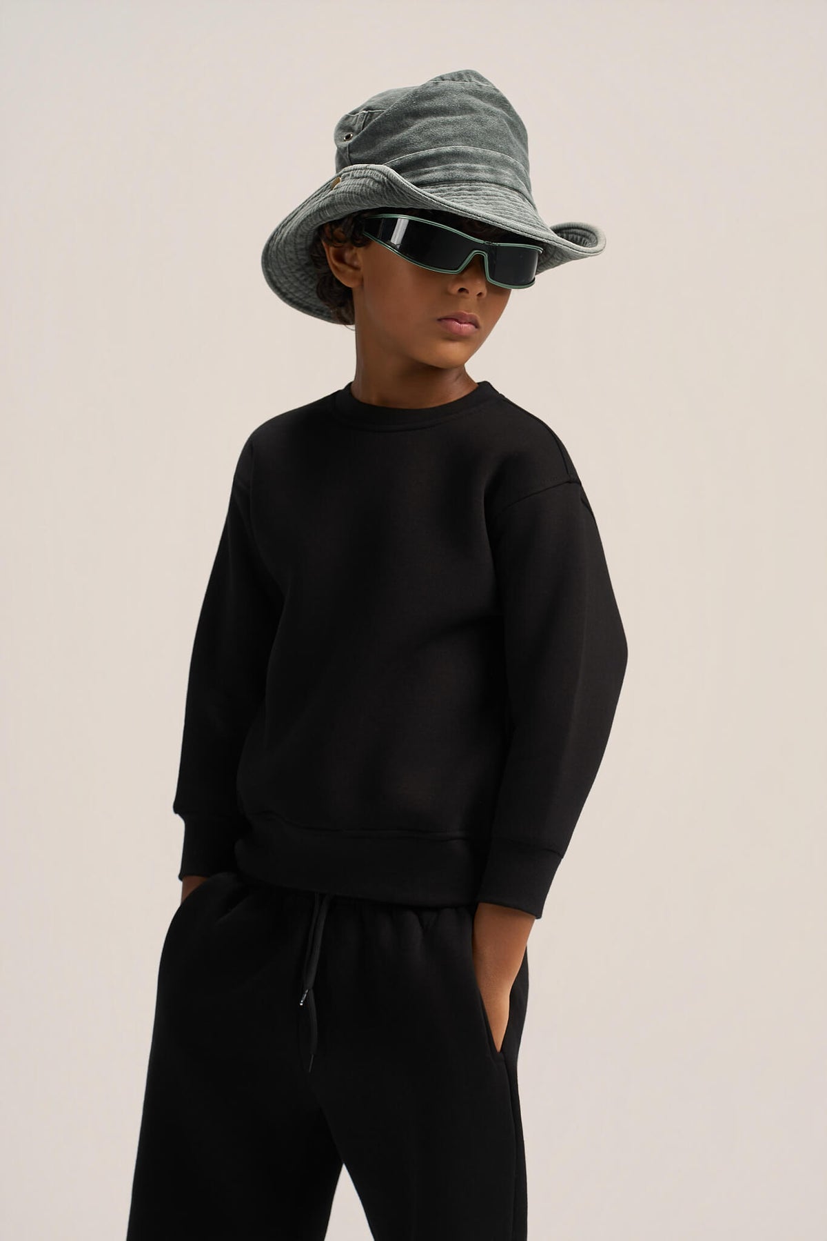 A child model wearing a black crewneck sweatshirt and black sweatpants, styled with a gray bucket hat and black sunglasses, against a plain light beige background.