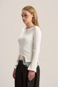 A woman modeling a fitted long-sleeve cream knit top with a sheer lace hem, paired with dark brown wide-leg pants, against a plain light beige studio background.