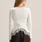 A woman shown from the back wearing a cream ribbed knit long-sleeve top with a silver-gray lace hem detail, paired with dark brown trousers, against a light beige studio background.