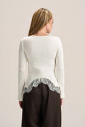 A woman shown from the back wearing a cream ribbed knit long-sleeve top with a silver-gray lace hem detail, paired with dark brown trousers, against a light beige studio background.