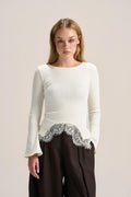 A woman modeling a cream long-sleeve knit top with a silver lace hem detail, paired with dark brown wide-leg trousers, against a solid light beige studio background.