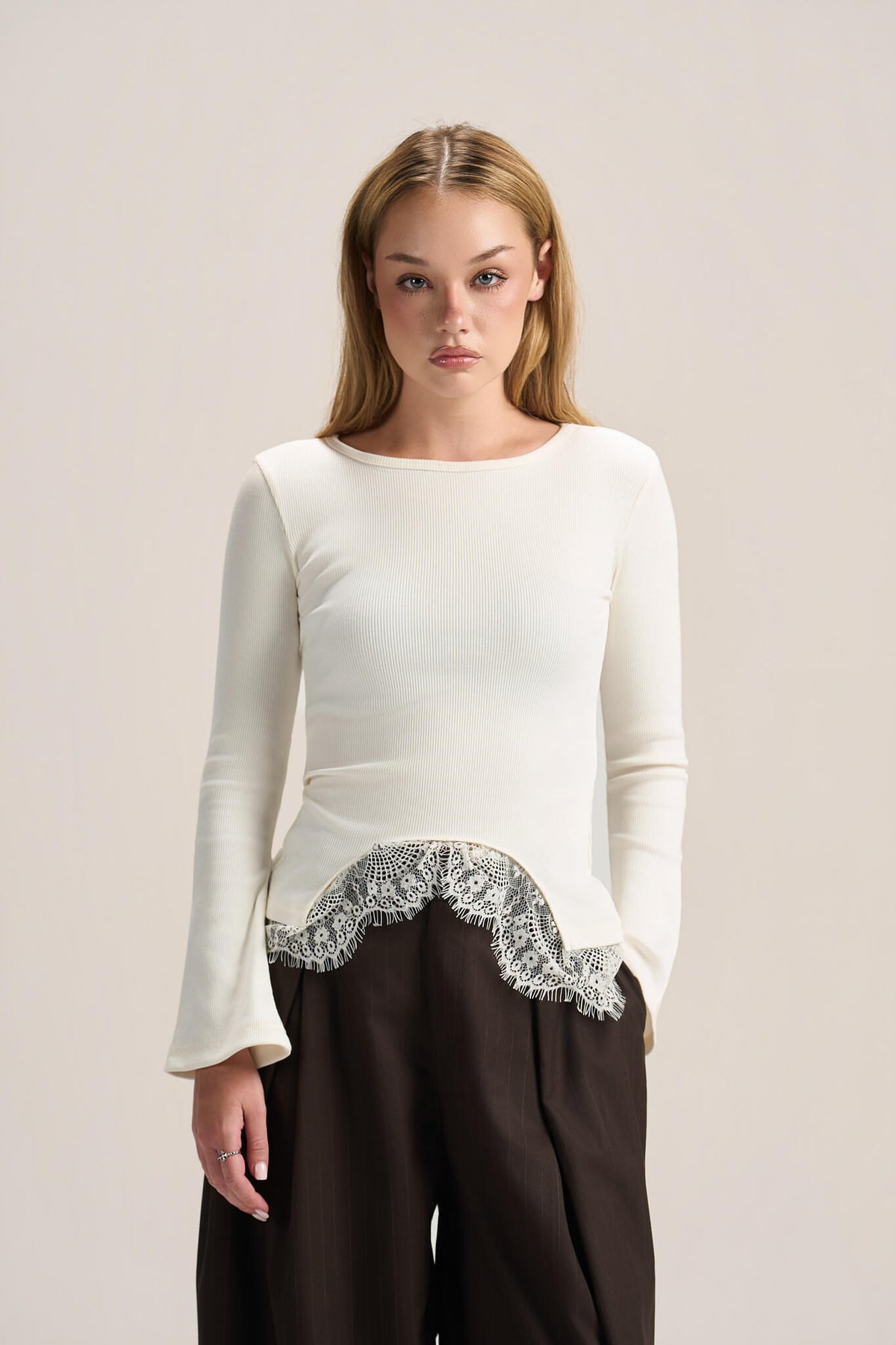 A woman modeling a cream long-sleeve knit top with a silver lace hem detail, paired with dark brown wide-leg trousers, against a solid light beige studio background.