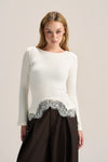 A woman modeling a white long-sleeve knit top with a scalloped silver lace hem, paired with a dark brown skirt, against a plain light beige studio background.