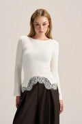 A woman modeling a white long-sleeve knit top with a scalloped silver lace hem, paired with a dark brown skirt, against a plain light beige studio background.
