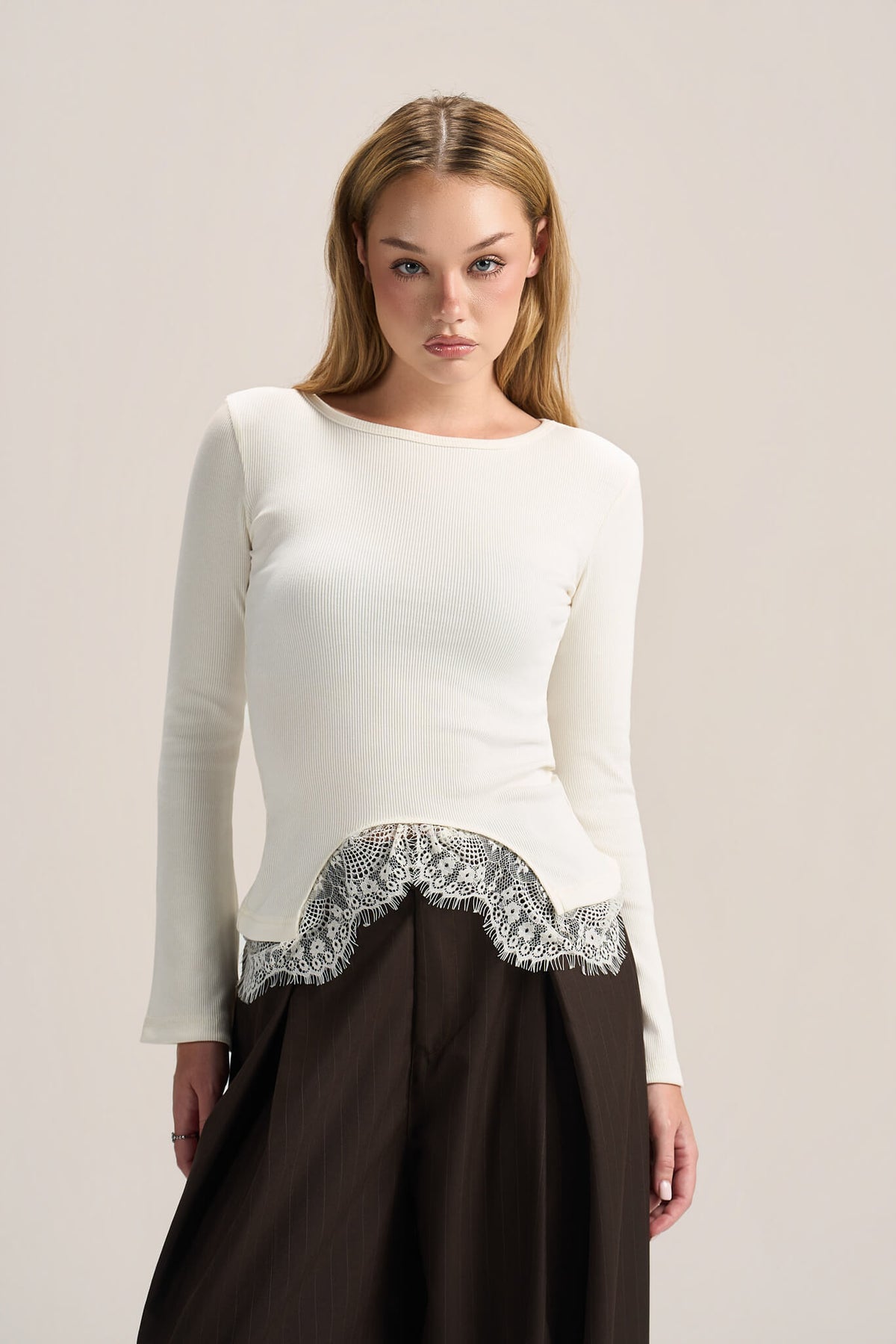 A woman modeling a white long-sleeve knit top with a scalloped silver lace hem, paired with a dark brown skirt, against a plain light beige studio background.