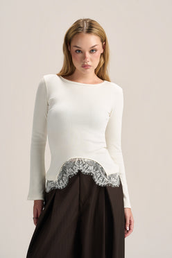 A woman modeling a white long-sleeve knit top with a scalloped silver lace hem, paired with a dark brown skirt, against a plain light beige studio background.