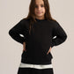 A young girl with long brown hair wearing a black crewneck sweatshirt and black pants, standing against a light beige studio background.