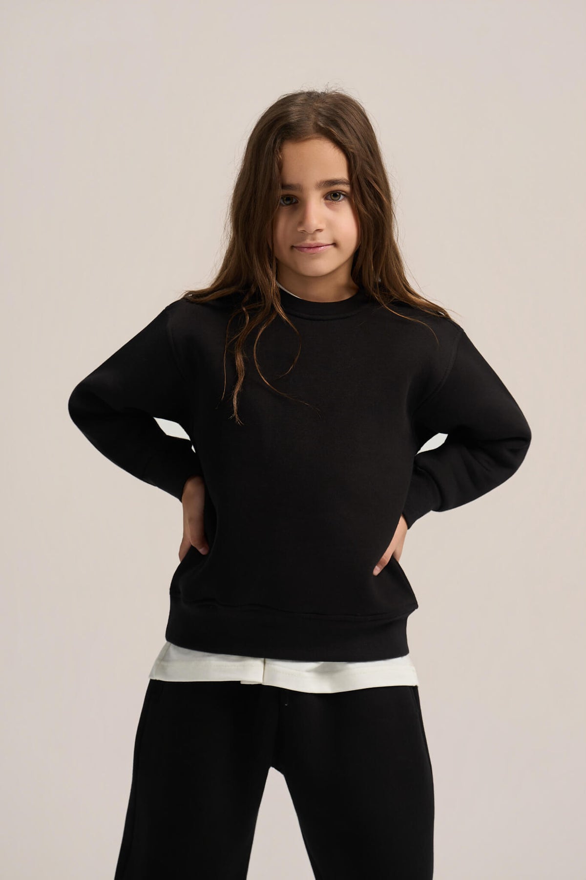 A young girl with long brown hair wearing a black crewneck sweatshirt and black pants, standing against a light beige studio background.