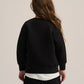 A model shown from the back wearing a black crewneck sweatshirt, a white undershirt, and black pants, set against a plain light beige studio background.