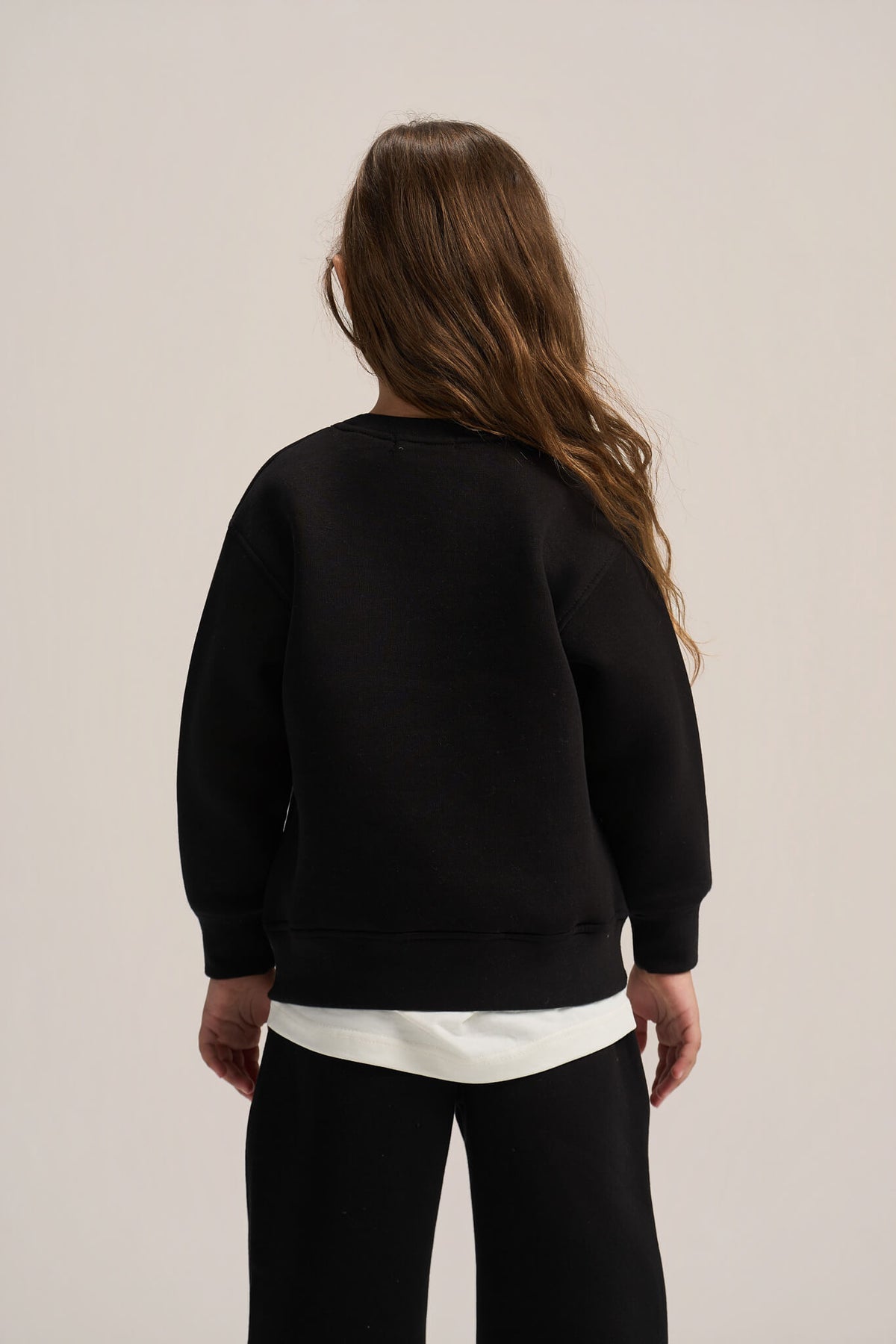 A model shown from the back wearing a black crewneck sweatshirt, a white undershirt, and black pants, set against a plain light beige studio background.