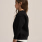 A child model shown in profile wearing a black crewneck sweatshirt over a white undershirt, set against a plain light beige studio background.