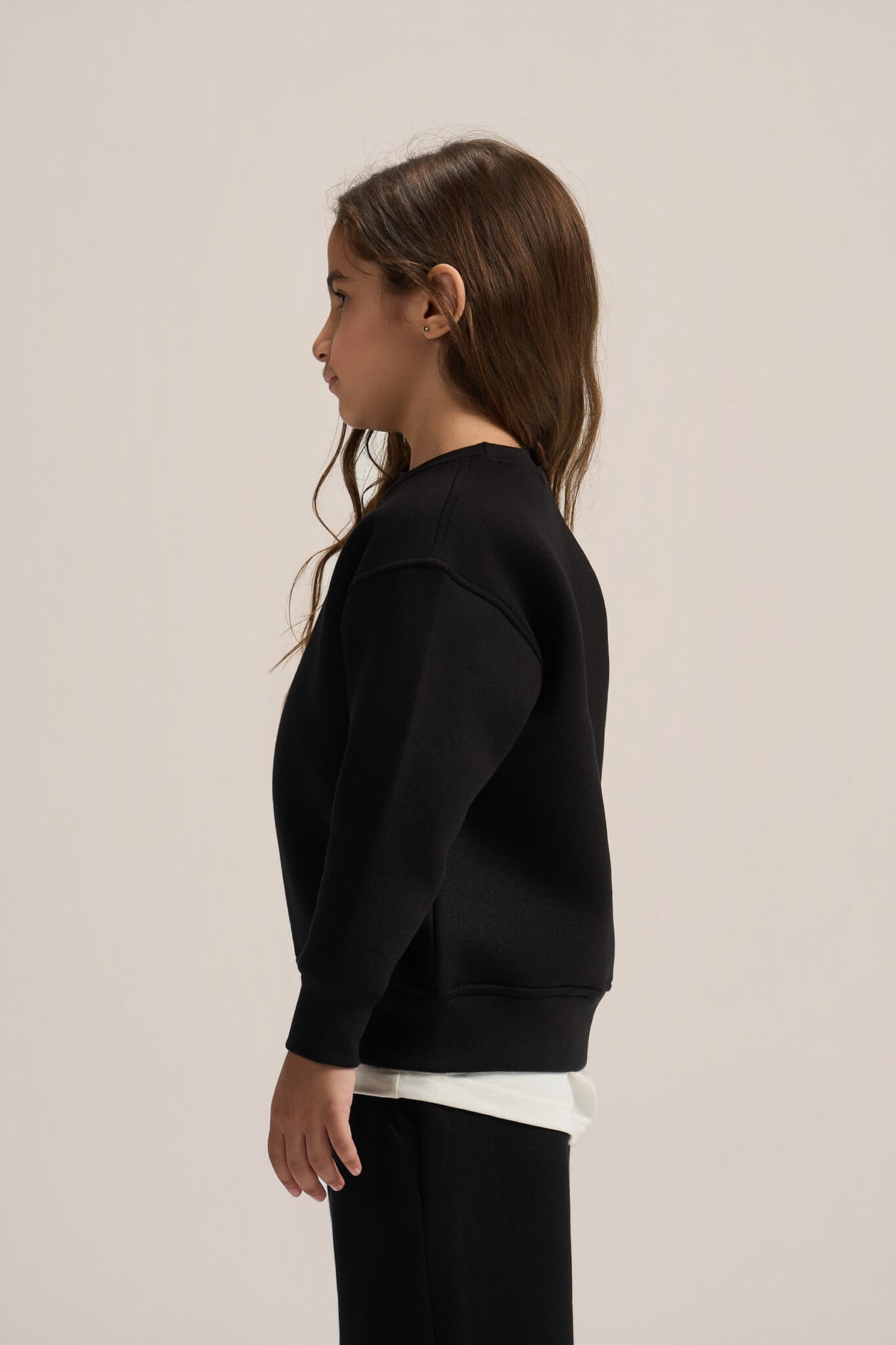 A child model shown in profile wearing a black crewneck sweatshirt over a white undershirt, set against a plain light beige studio background.