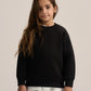 A young girl with long brown hair wearing a black crewneck sweatshirt over a white t-shirt and black pants, standing against a plain light beige studio background.