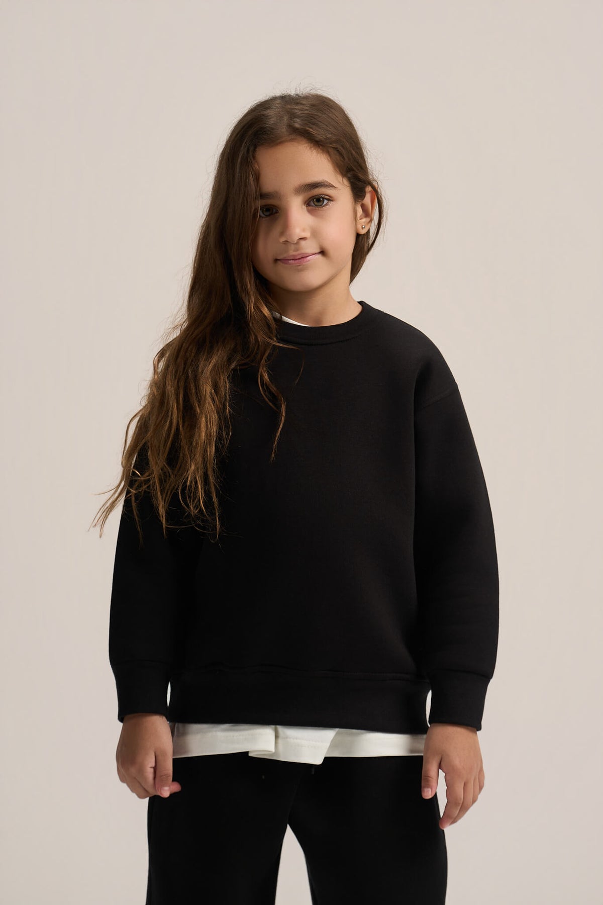 A young girl with long brown hair wearing a black crewneck sweatshirt over a white t-shirt and black pants, standing against a plain light beige studio background.