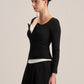 A woman modeling a black long-sleeve knit top with a unique asymmetrical zipper neckline, layered over a white top and paired with a black skirt, against a plain light gray background.