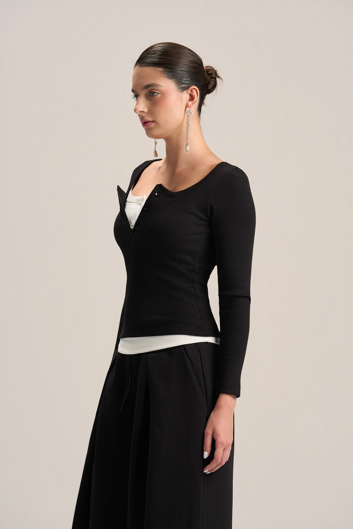 A woman modeling a black long-sleeve knit top with a unique asymmetrical zipper neckline, layered over a white top and paired with a black skirt, against a plain light gray background.