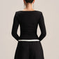 A woman viewed from the back wearing a fitted black long-sleeve top with a wide boat neckline and black wide-leg pants, against a plain light gray studio background.