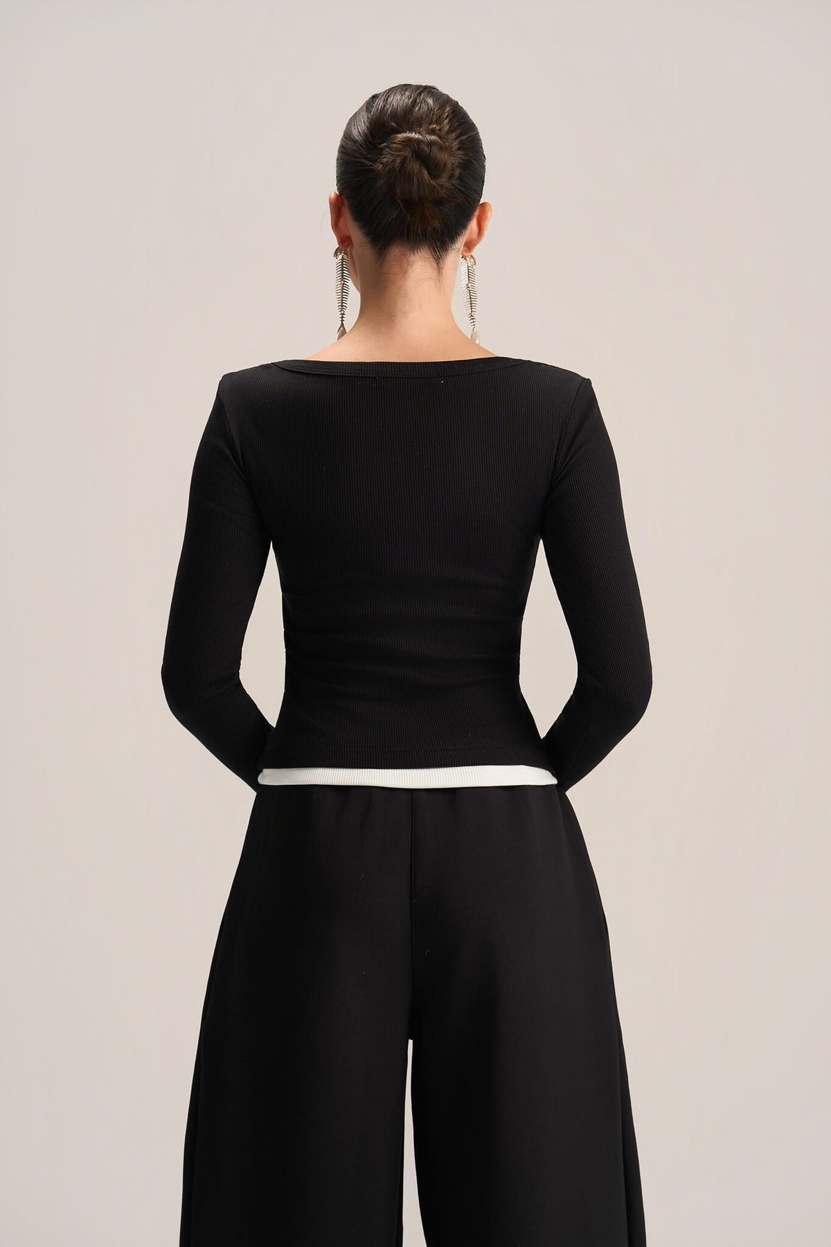 A woman viewed from the back wearing a fitted black long-sleeve top with a wide boat neckline and black wide-leg pants, against a plain light gray studio background.