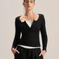 A model wearing a black long-sleeve knit top with a unique scalloped neckline over a white top, paired with black drawstring trousers, against a solid light beige background.