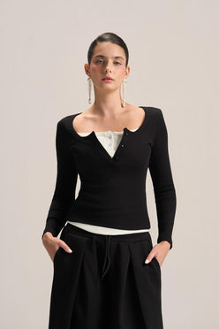 A model wearing a black long-sleeve knit top with a unique scalloped neckline over a white top, paired with black drawstring trousers, against a solid light beige background.