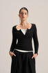 A model wearing a black long-sleeve knit top with a unique scalloped neckline over a white top, paired with black drawstring trousers, against a solid light beige background.