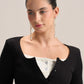 A woman with her hair pulled back wears a black ribbed long-sleeve top over a white henley-style undershirt, accessorized with long silver dangle earrings, against a plain light beige studio background.