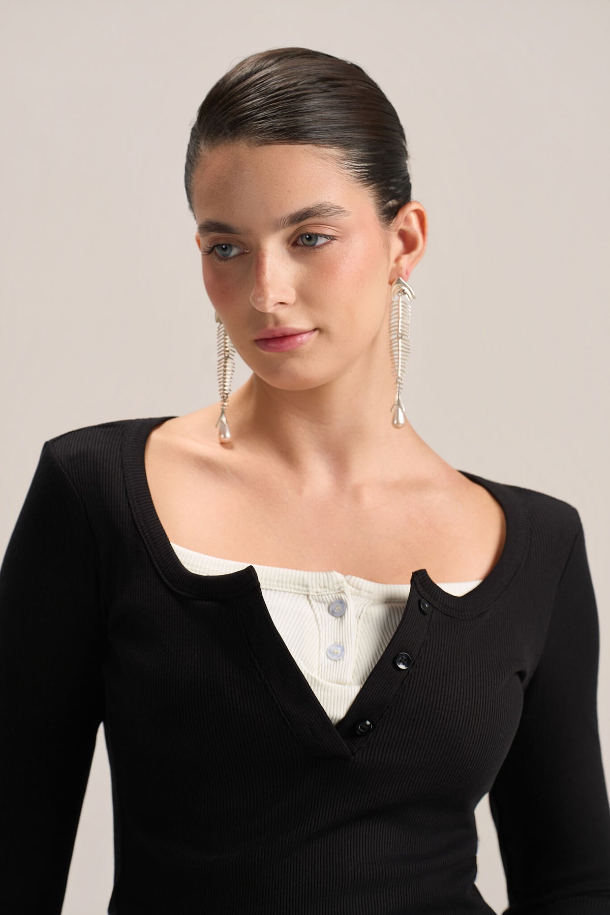 A woman with her hair pulled back wears a black ribbed long-sleeve top over a white henley-style undershirt, accessorized with long silver dangle earrings, against a plain light beige studio background.