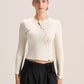A model wearing a cream ribbed long-sleeve button-front crop top and black pleated trousers, standing against a solid light beige background.