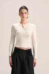 A model wearing a cream ribbed long-sleeve button-front crop top and black pleated trousers, standing against a solid light beige background.