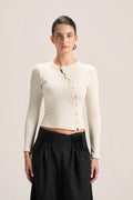 A model wearing a cream ribbed long-sleeve button-front crop top and black pleated trousers, standing against a solid light beige background.
