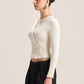 A woman in side profile wearing a fitted, long-sleeve cream knit crop top with a buttoned front and black wide-leg pants, photographed against a plain light beige studio background.