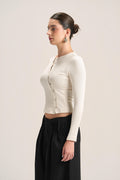 A woman in side profile wearing a fitted, long-sleeve cream knit crop top with a buttoned front and black wide-leg pants, photographed against a plain light beige studio background.