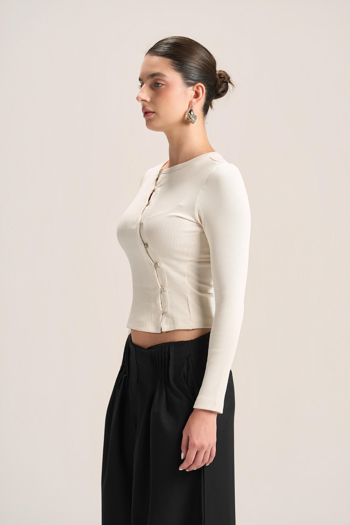 A woman in side profile wearing a fitted, long-sleeve cream knit crop top with a buttoned front and black wide-leg pants, photographed against a plain light beige studio background.