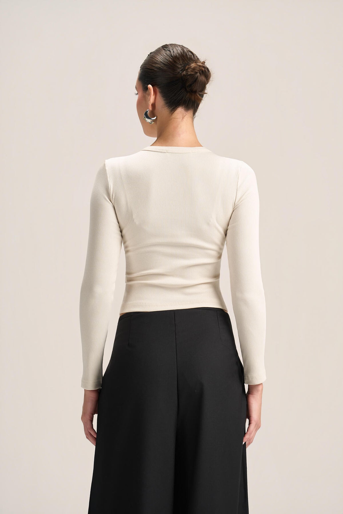 A woman shown from the back wearing a fitted long-sleeve cream top and black wide-leg pants against a plain light beige studio background.