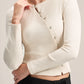 A woman wearing a ribbed cream-colored long-sleeve cardigan with a buttoned front and a small bow detail at the neckline, paired with black pleated trousers.