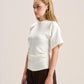 A blonde model wearing a white short-sleeve top with structured shoulders and gathered detailing at the waist, paired with dark brown wide-leg pants, photographed against a plain light beige studio background.