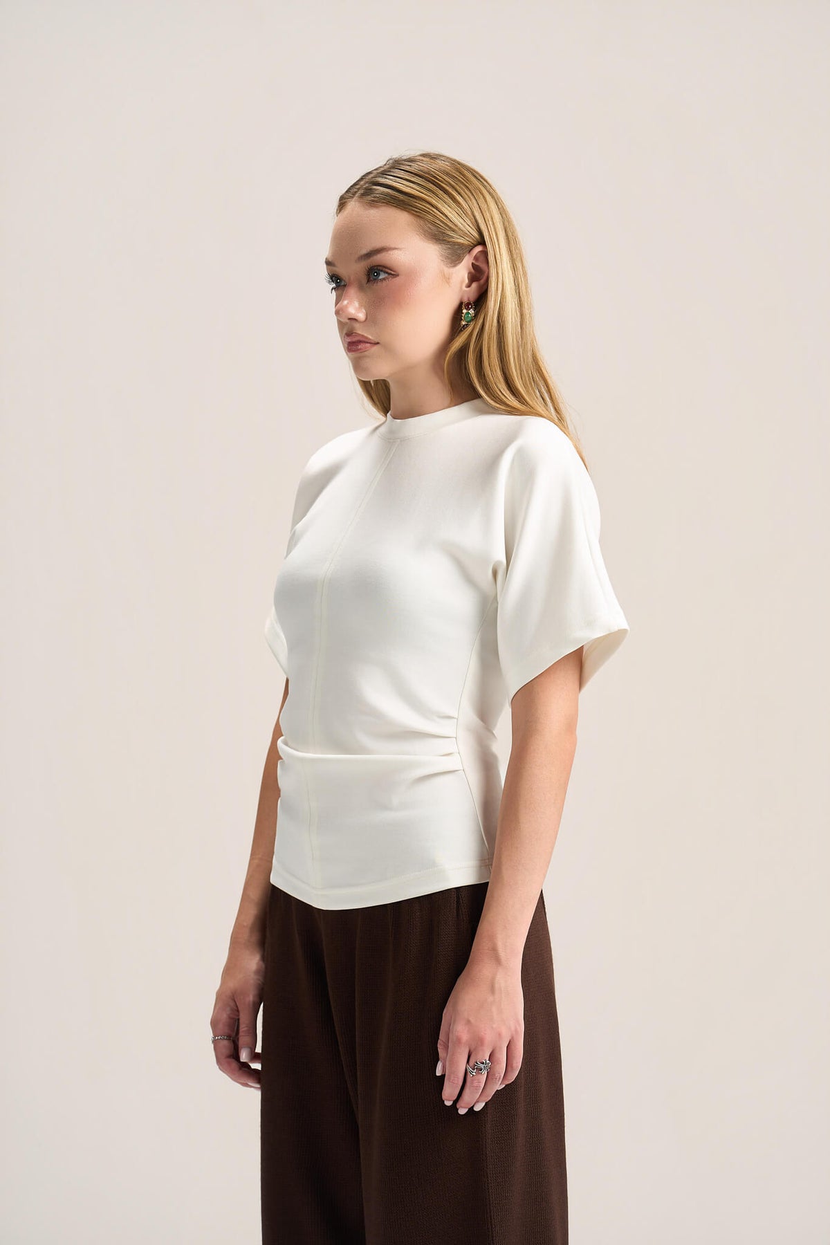 A blonde model wearing a white short-sleeve top with structured shoulders and gathered detailing at the waist, paired with dark brown wide-leg pants, photographed against a plain light beige studio background.