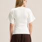 A woman viewed from the back wearing a cream-colored short-sleeve knit top and dark brown wide-leg trousers, set against a plain light beige studio background.