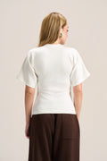 A woman viewed from the back wearing a cream-colored short-sleeve knit top and dark brown wide-leg trousers, set against a plain light beige studio background.