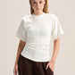 A woman modeling a short-sleeve white mock-neck top with a unique gathered waist, paired with dark brown wide-leg pants, against a solid light beige background.