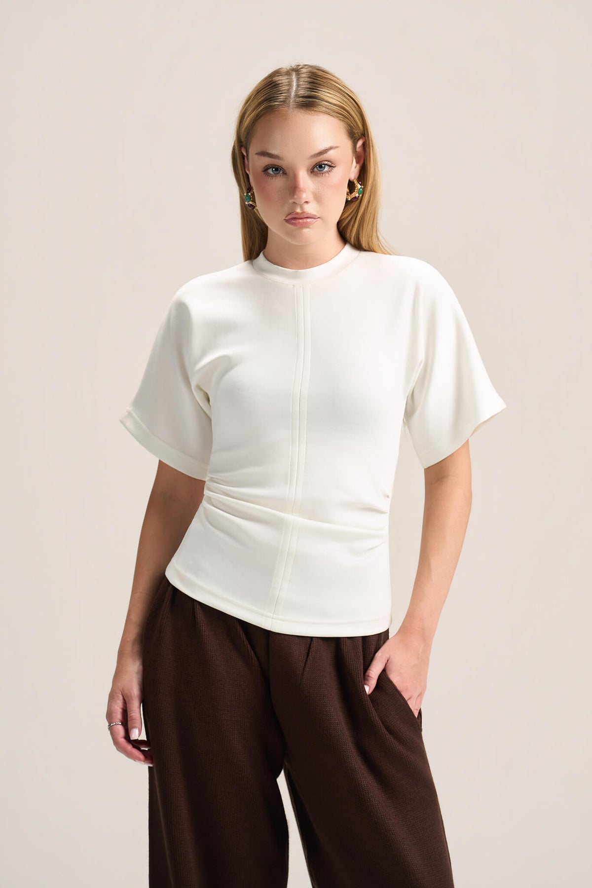A woman modeling a short-sleeve white mock-neck top with a unique gathered waist, paired with dark brown wide-leg pants, against a solid light beige background.