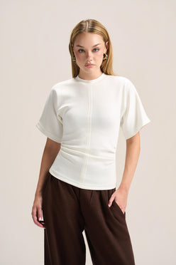 A woman modeling a short-sleeve white mock-neck top with a unique gathered waist, paired with dark brown wide-leg pants, against a solid light beige background.