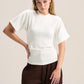 A woman modeling a white short-sleeve mock-neck knit top with a structured waist, paired with dark brown wide-leg trousers, against a solid light beige background.