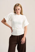 A woman modeling a white short-sleeve mock-neck knit top with a structured waist, paired with dark brown wide-leg trousers, against a solid light beige background.