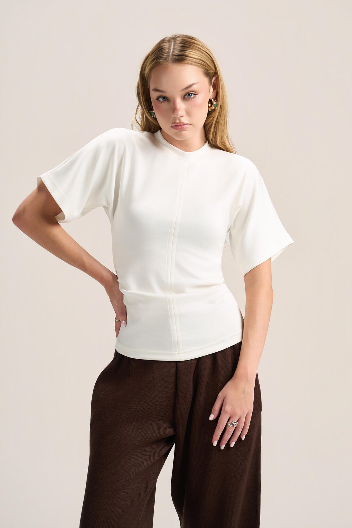 A woman modeling a white short-sleeve mock-neck knit top with a structured waist, paired with dark brown wide-leg trousers, against a solid light beige background.