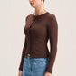 A woman wearing a fitted long-sleeve brown cardigan with front buttons and blue jeans, standing against a plain light beige studio background.