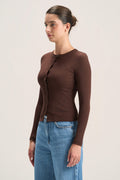 A woman wearing a fitted long-sleeve brown cardigan with front buttons and blue jeans, standing against a plain light beige studio background.