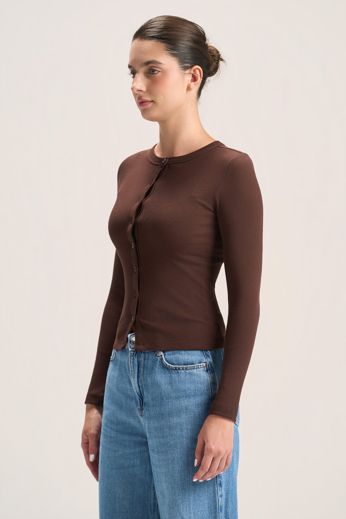 A woman wearing a fitted long-sleeve brown cardigan with front buttons and blue jeans, standing against a plain light beige studio background.