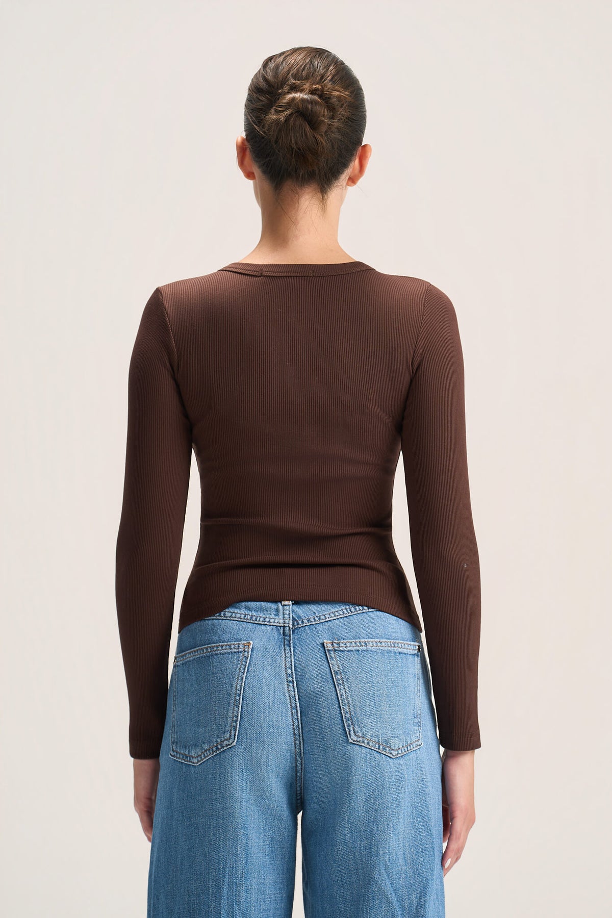 A woman viewed from the back wearing a fitted brown long-sleeve top and light blue jeans against a plain light background.